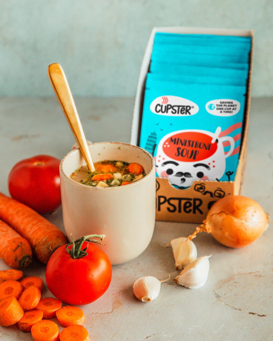 A box of Cupster minestrone soup packet stands behind a white mug filled with soup and a wooden spoon, surrounded by fresh ingredients including tomatoes, carrots, garlic, and an onion on a light stone surface.