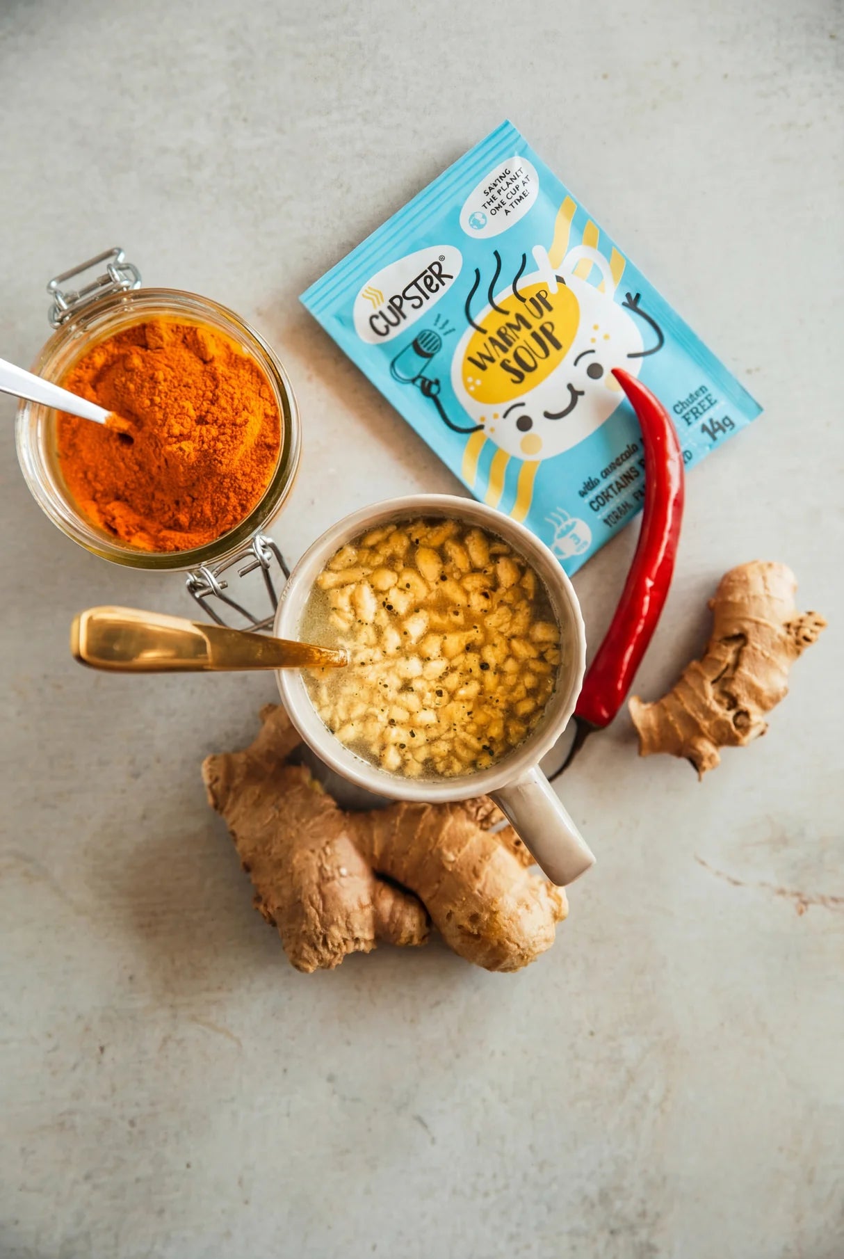 Cupster instant turkish red lentil creamsoup snack mix packet surrounded by its ingredients: a glass jar of orange-red powder, a bowl of yellow flakes, ginger roots, and a red chili pepper on a light gray textured background.