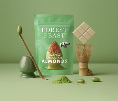 Forest Feast Matcha Chocolate Almonds in a mint green stand-up pouch, styled with a bamboo whisk, matcha powder, green chocolate covered almonds, and a white chocolate bar on a matching sage green background.