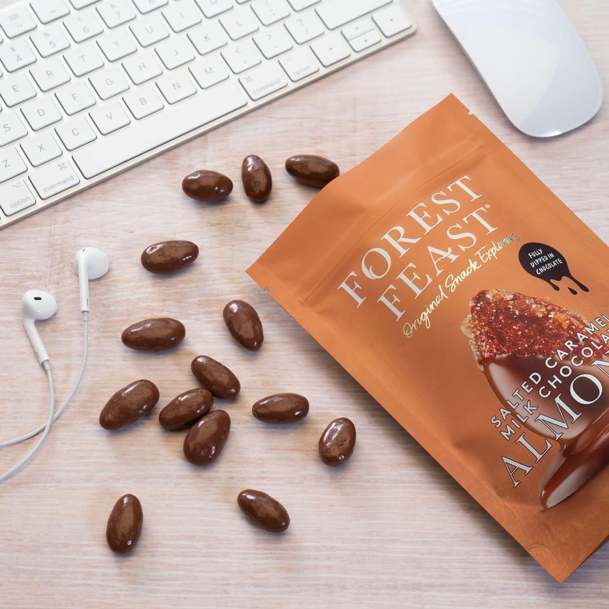 Opened pack of Forest Feast Salted Caramel Milk Chocolate Almonds Chocolate Nuts 120g Almonds scattered around it. A white keyboard, mouse, and earbuds are on the desk