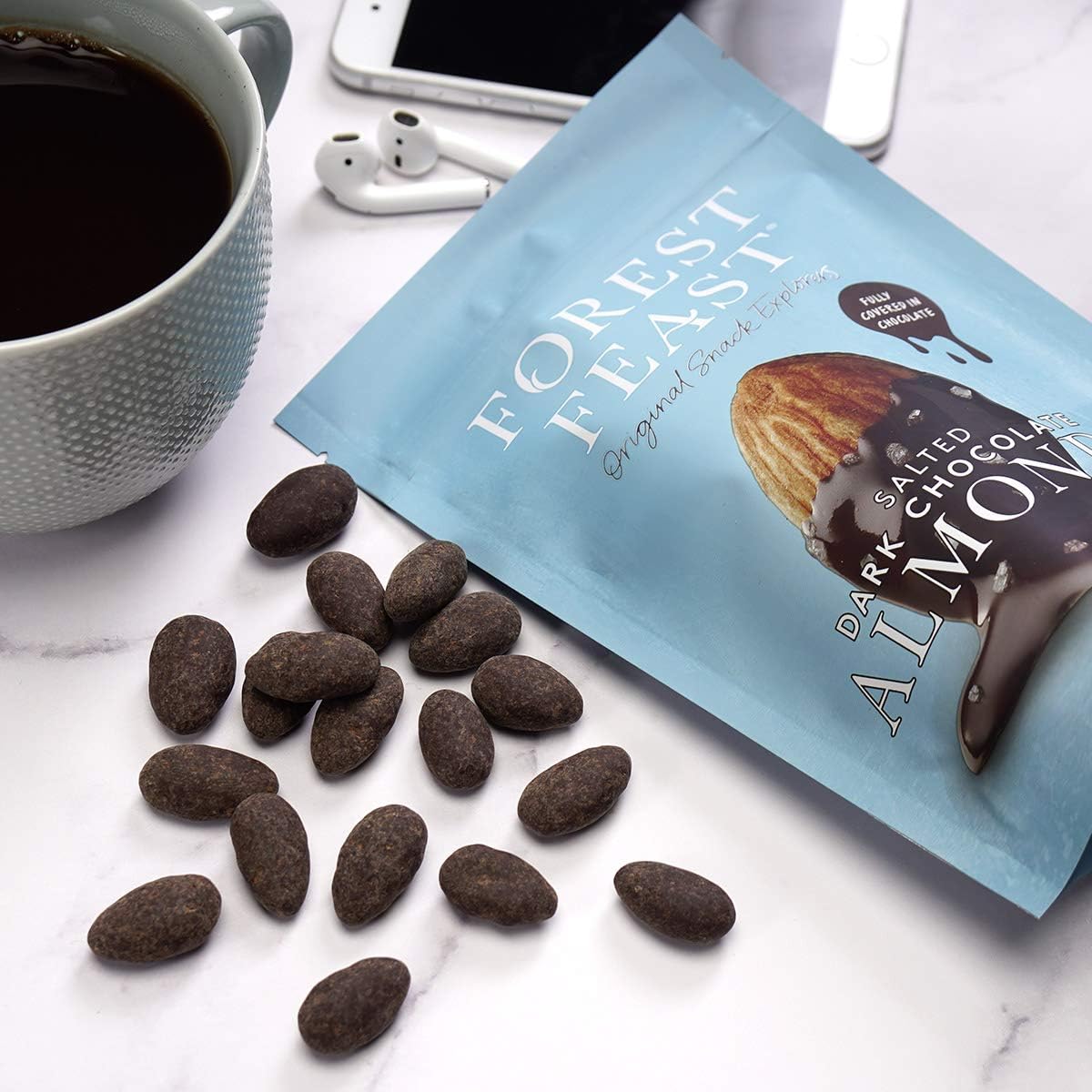 Packaged dark chocolate almonds with a cup of coffee and phone on a marble surface