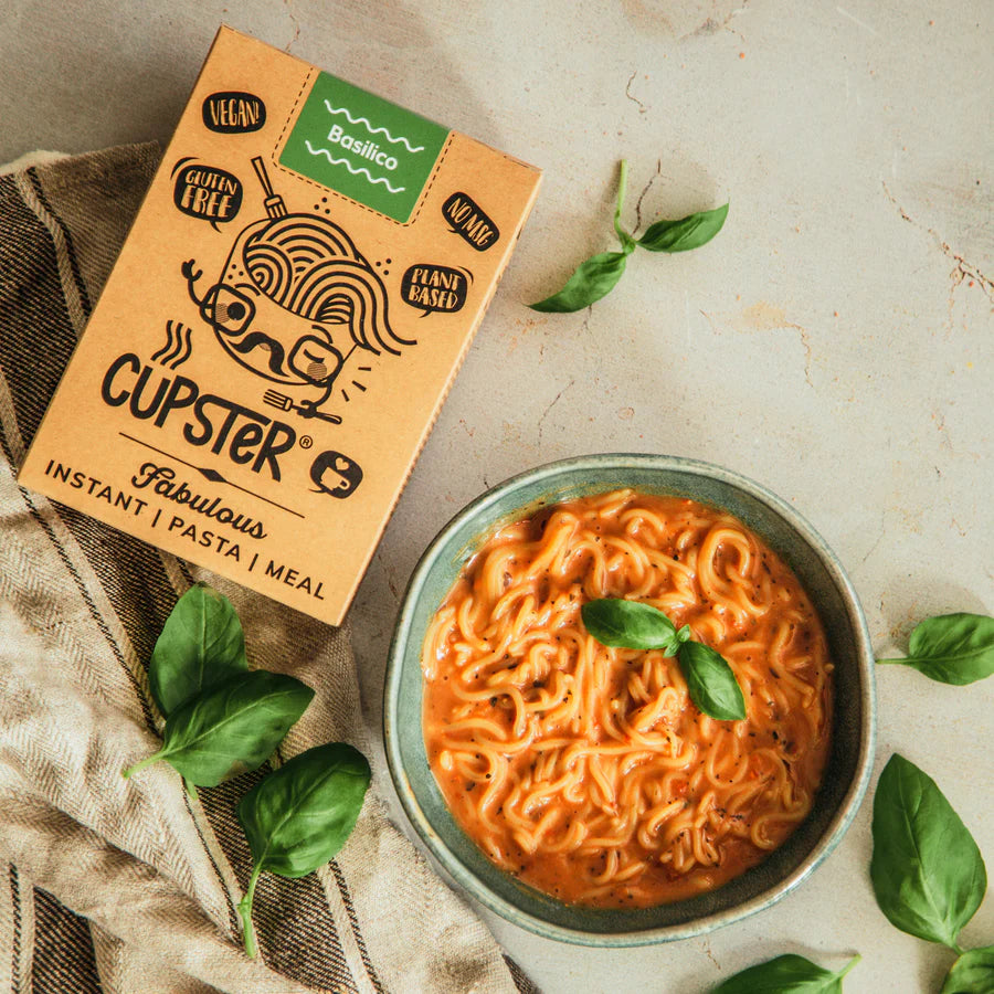 Cupster Fabulous Instant Pasta & Meal box shown next to a bowl of prepared orange pasta with basil leaves on a light stone surface with scattered basil.