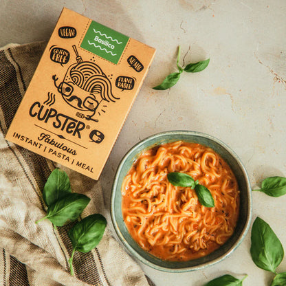 Cupster Fabulous Instant Pasta & Meal box shown next to a bowl of prepared orange pasta with basil leaves on a light stone surface with scattered basil.