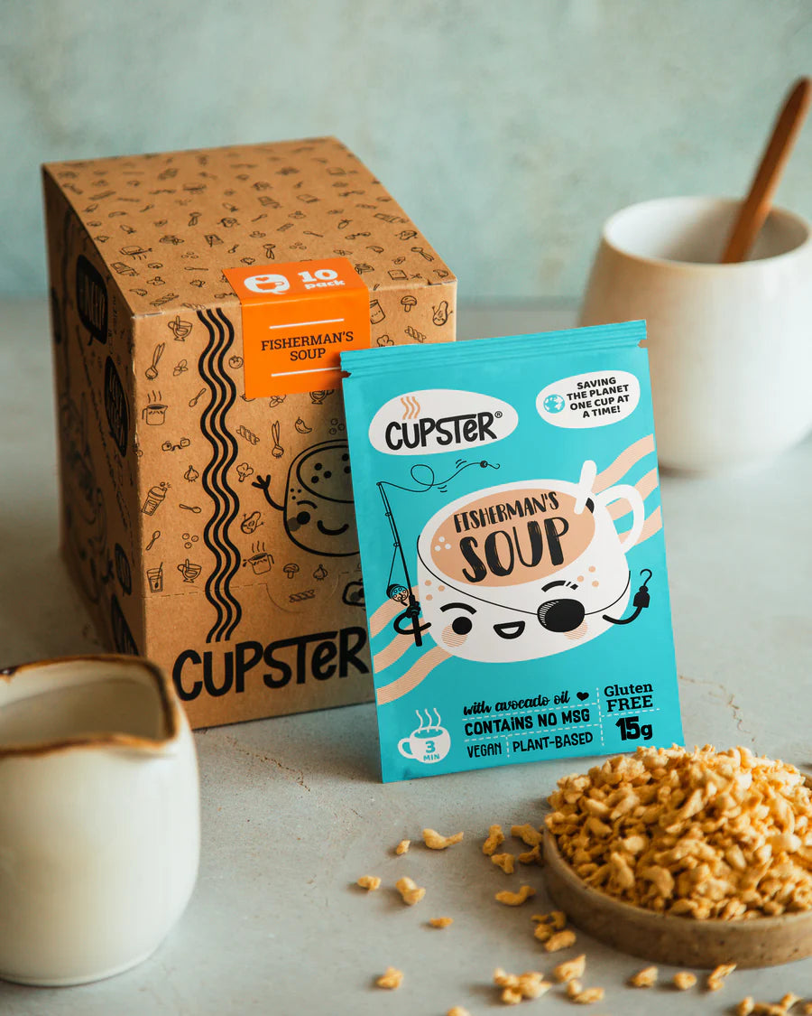Cupster Fisherman's Soup packet in front of its brown cardboard 10 pack box, styled on a light gray surface with a bowl of rice and white ceramic cups.