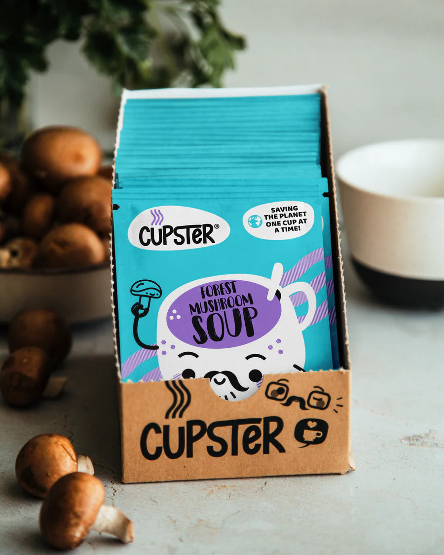 A box of 10 Cupster forest mushroom soup packets, featuring bright blue and brown packaging with a cartoon mushroom character, styled on a light gray surface with mushrooms and a bowl in the background.