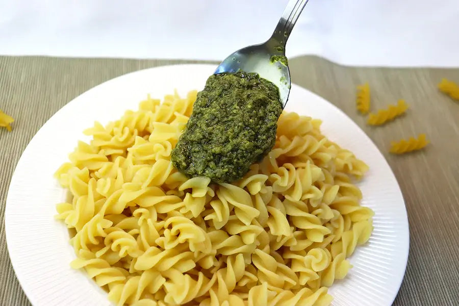 A close-up of a white plate filled with rotini pasta, topped with a spoonful of green pesto sauce, set on a beige placemat with a few loose pasta pieces nearby.