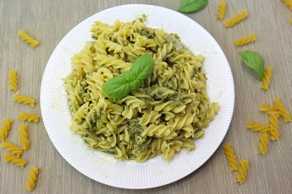 A white plate of rotini pasta tossed with green pesto sauce and garnished with a fresh basil leaf, set on a beige woven placemat with scattered pasta pieces around it.