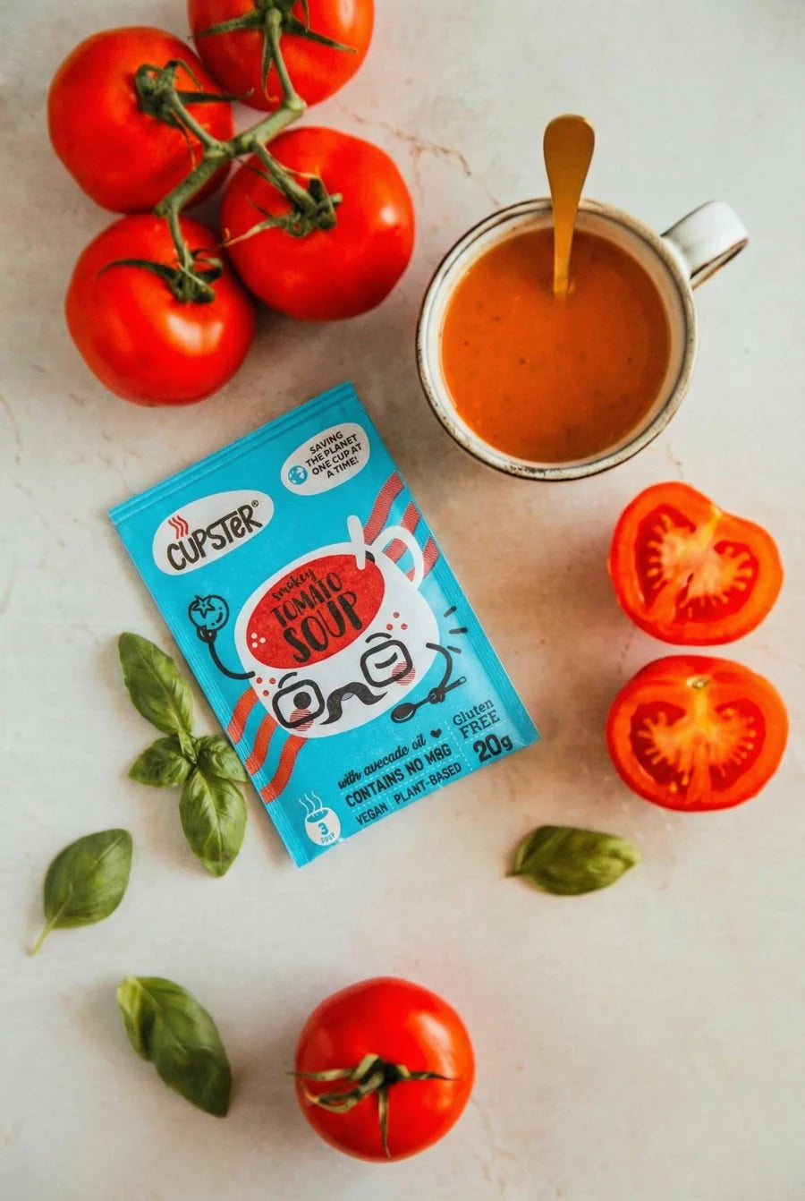 Cupster instant smokey tomato soup pack with a cup of soup, tomatoes, and basil leaves on a light surface