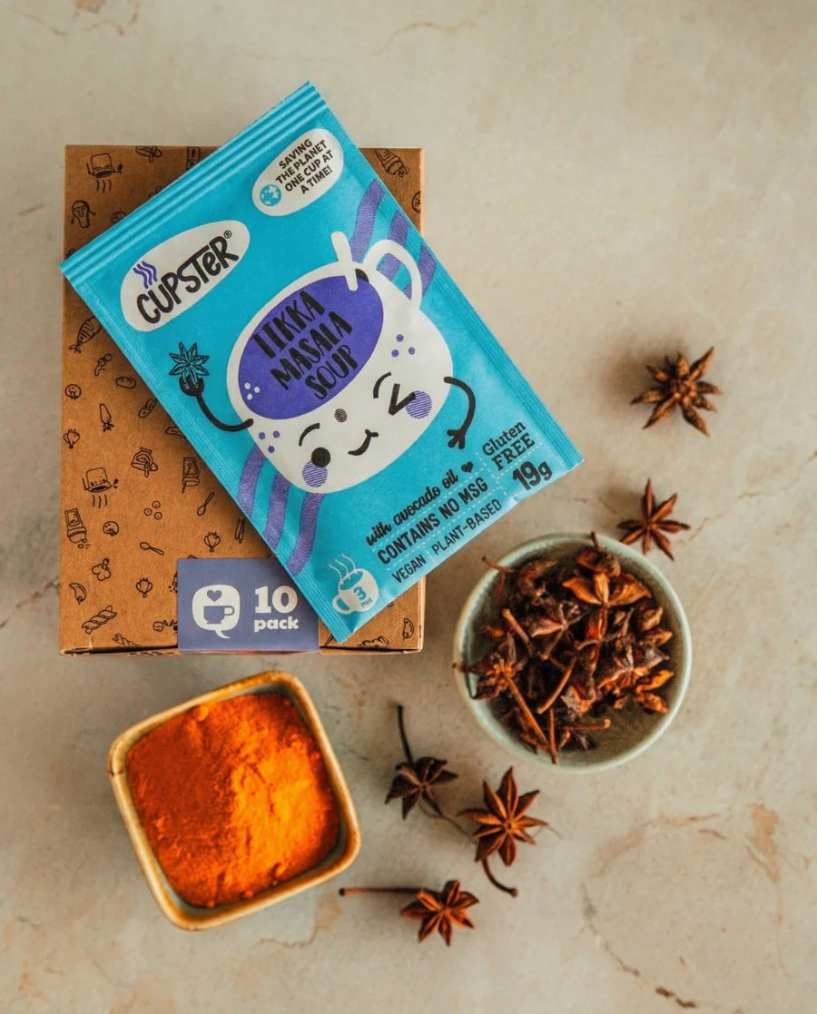 Cupster instant tikka masala soup 10 pack displayed on top of a brown cardboard box with star anise and bowls of orange powder and spices on a beige stone surface.