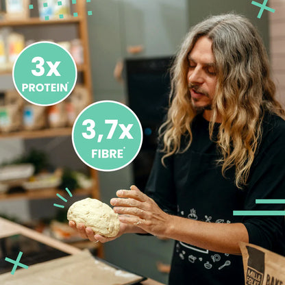 A man with long wavy gray hair kneads a ball of dough in a kitchen setting, wearing a black t-shirt. Two mint green circular text overlays highlight the product's nutritional benefits: '3x PROTEIN' and '3,7x FIBRE'.
