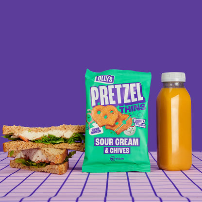 Olly's - Sour Cream & Chives Pretzels with a sandwich and juice bottle on a purple grid background